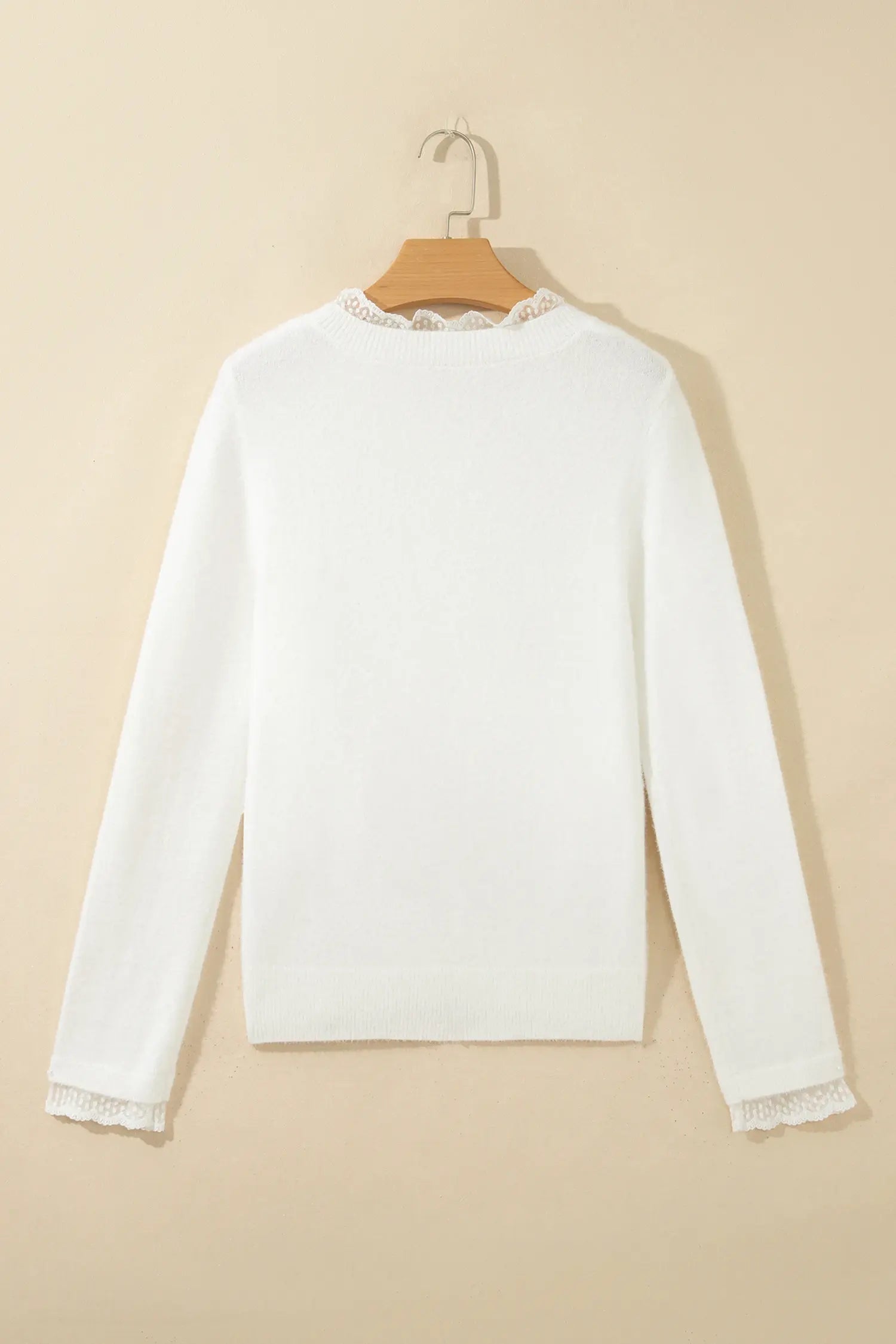 A white long-sleeve knit sweater hanging on a wooden hanger against a warm beige background. The sweater features delicate lace trim detailing at the round neckline and the cuffs.