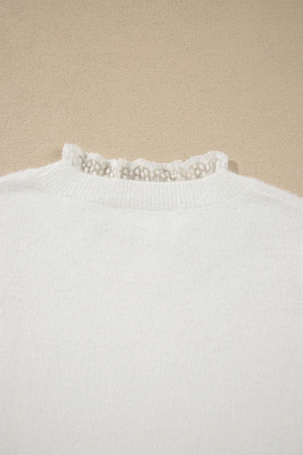 Close-up of a white knit sweater with a delicate lace ruffle trim at the crew neck, set against a neutral beige background.