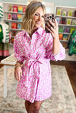 A woman takes a mirror selfie in a boutique while wearing a pink and white leopard-print shirt dress with a tie waist. The background shows clothing racks and shelves with colorful items.