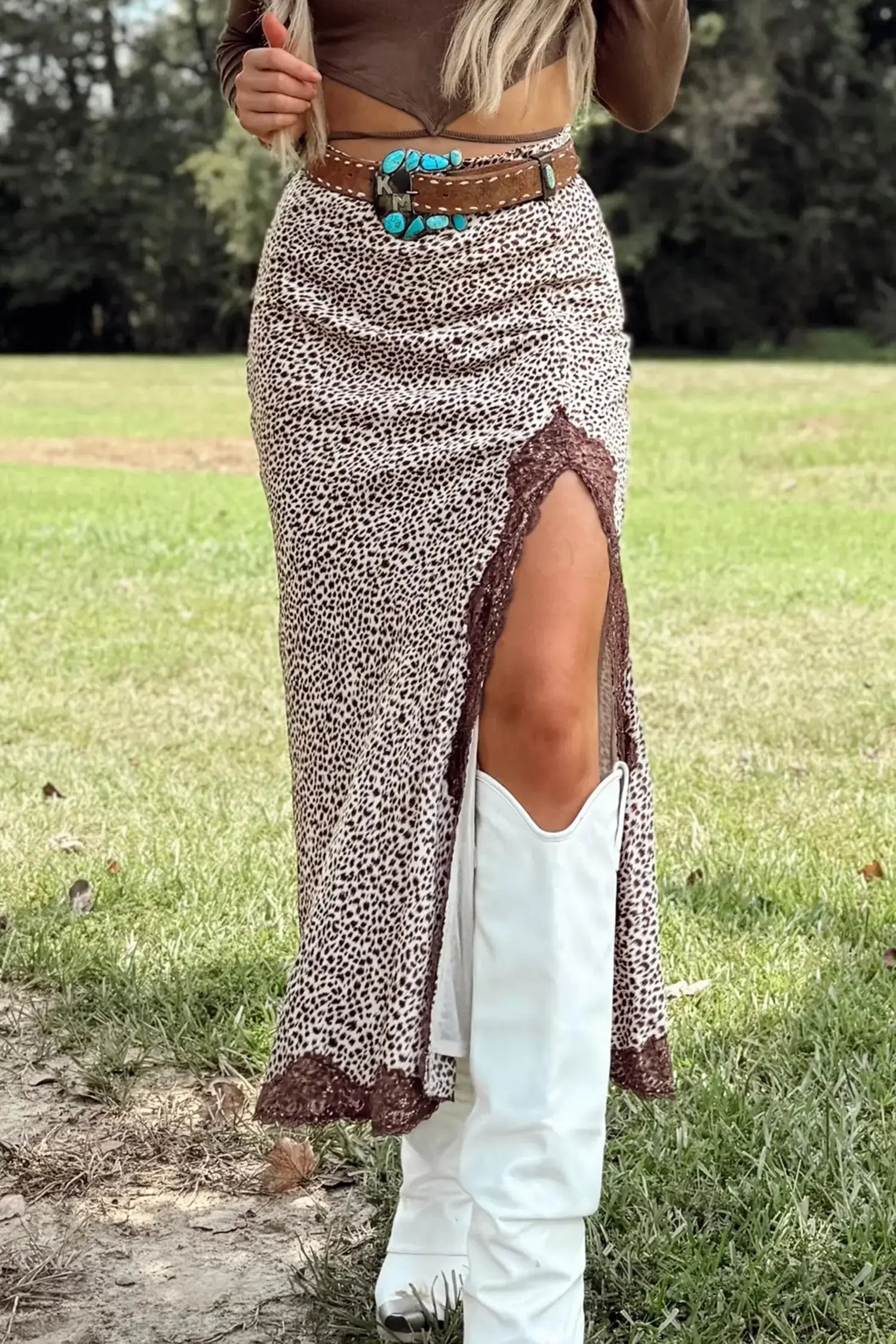 A woman outdoors wearing a brown and beige leopard-print maxi skirt with a high side slit, a brown belt with a turquoise concho, and white cowboy boots.