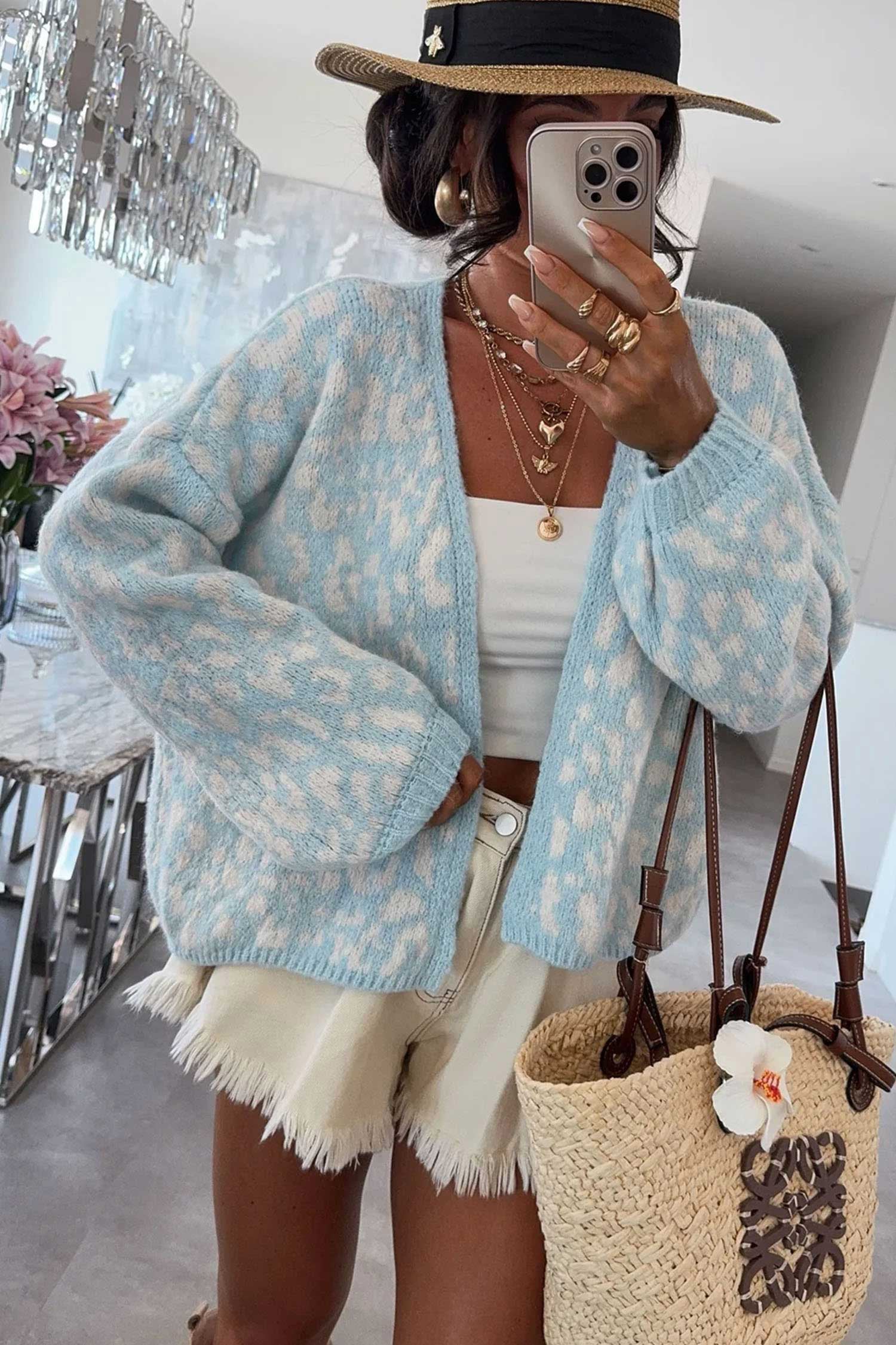 A woman takes a mirror selfie wearing a light blue and white patterned knit cardigan, a white tank top, frayed cream shorts, a black and beige hat, and carrying a woven straw tote bag with brown handles in a bright indoor setting.