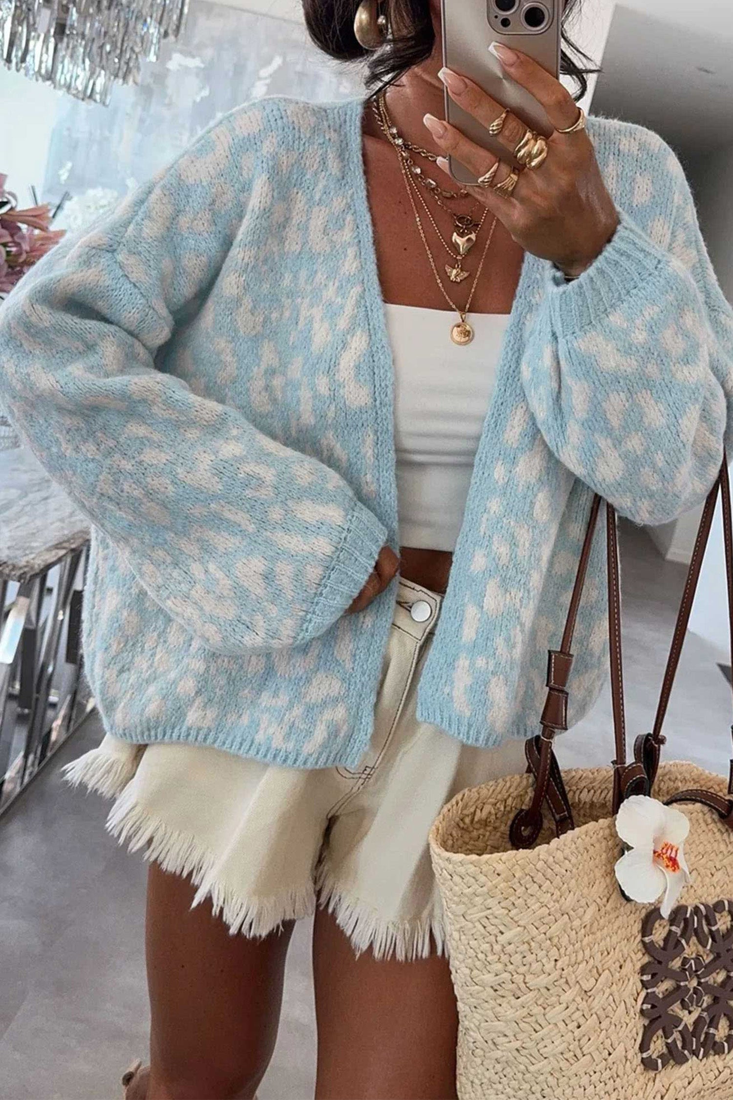 A woman takes a mirror selfie wearing a light blue and white patterned knit cardigan, a white tank top, and cream frayed denim shorts. She is accessorized with layered gold necklaces and rings, and carries a woven straw tote bag with brown leather handles.