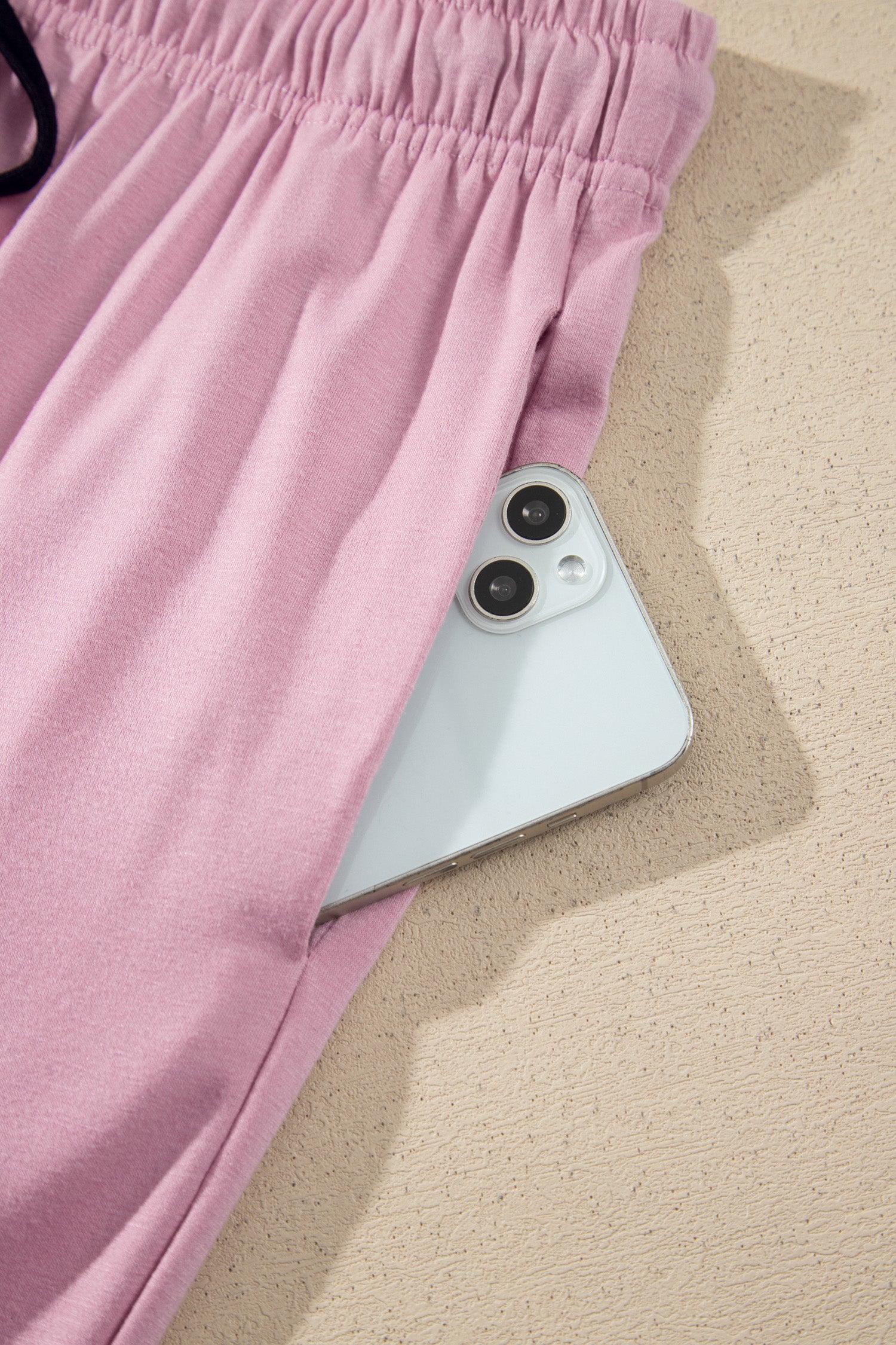 A close-up of light pink drawstring shorts with a side pocket, shown with a white smartphone tucked inside the pocket on a textured beige surface.
