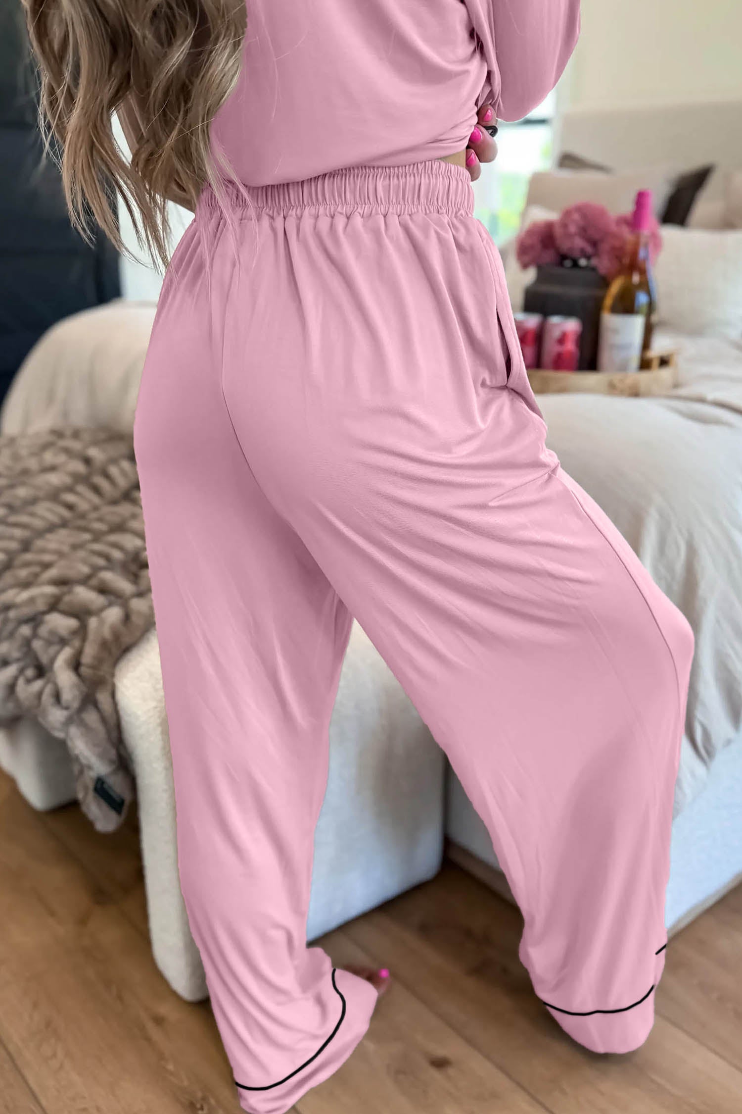 A person wearing a light pink two-piece loungewear set with matching long-sleeve top and wide-leg pants, standing in a bedroom setting.