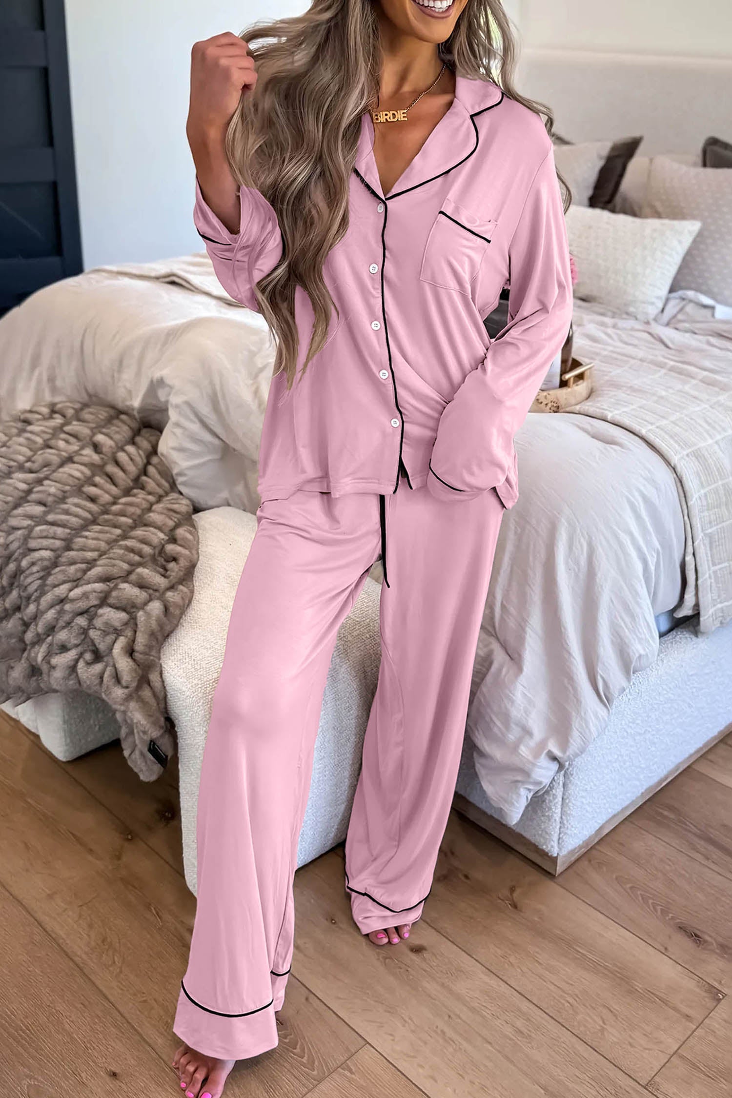 A woman models a two-piece pink pajama set with black piping in a bedroom setting. The set includes a long-sleeve button-up top and matching wide-leg pants.