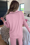 A woman viewed from behind wearing a light pink long-sleeve pajama set with thin black piping trim on the collar, standing in a bedroom.