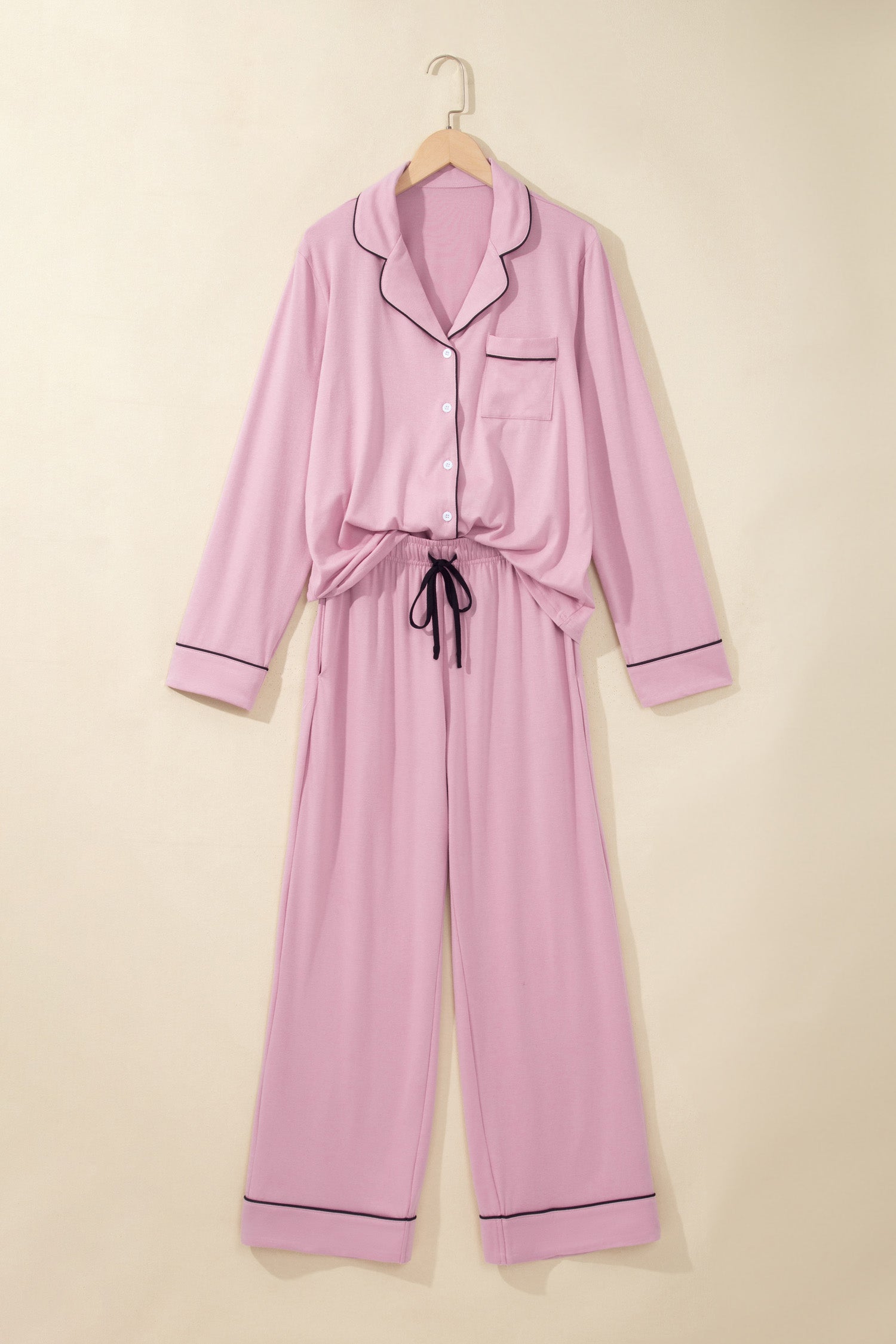 A light pink long-sleeve pajama set with dark piping trim, featuring a button-up front, chest pocket, and self-tie belt, hanging on a wooden hanger against a solid beige background.