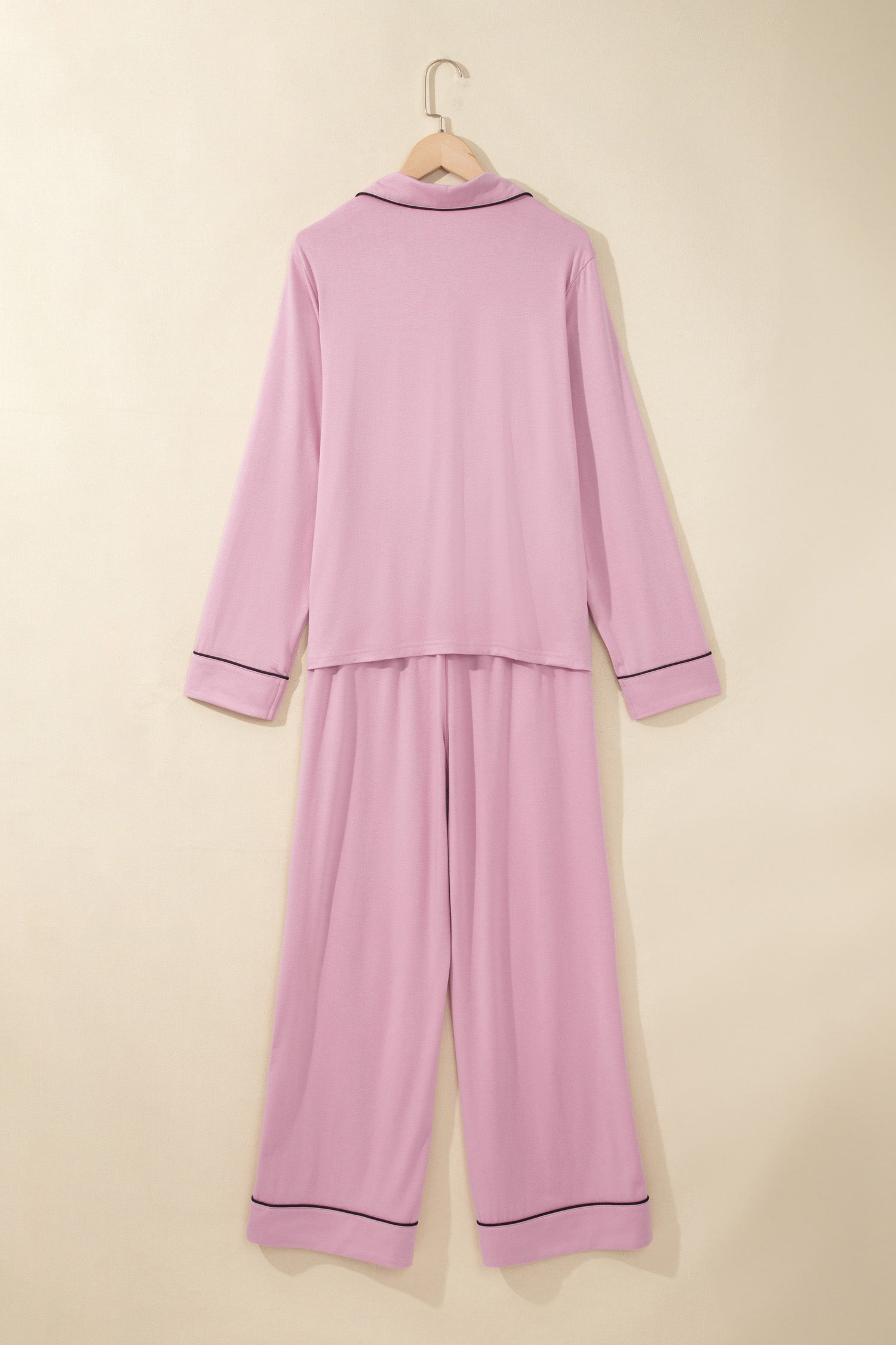 A two-piece long-sleeve pajama set in light pink, hanging on a wooden hanger against a solid beige background. The set features a matching top and wide-leg pants with thin dark piping trim along the collar, cuffs, and hem.