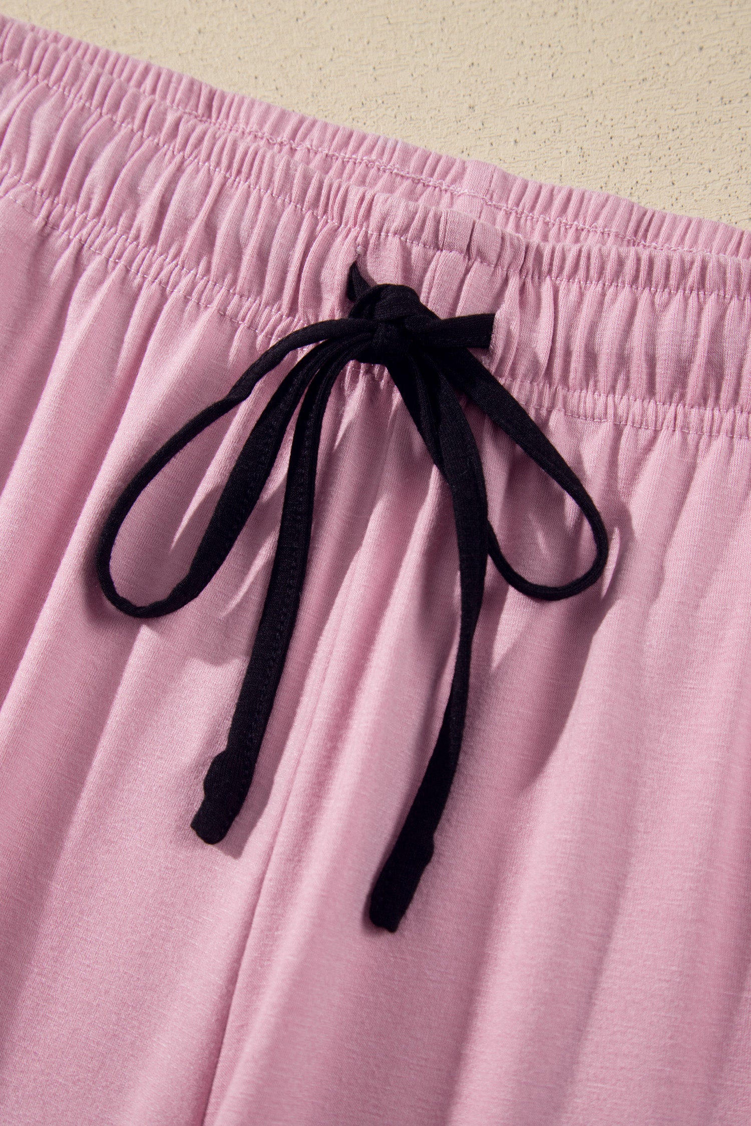 Close-up of a pair of light pink lounge pants with a black drawstring at the elastic waistband, set against a neutral beige background.