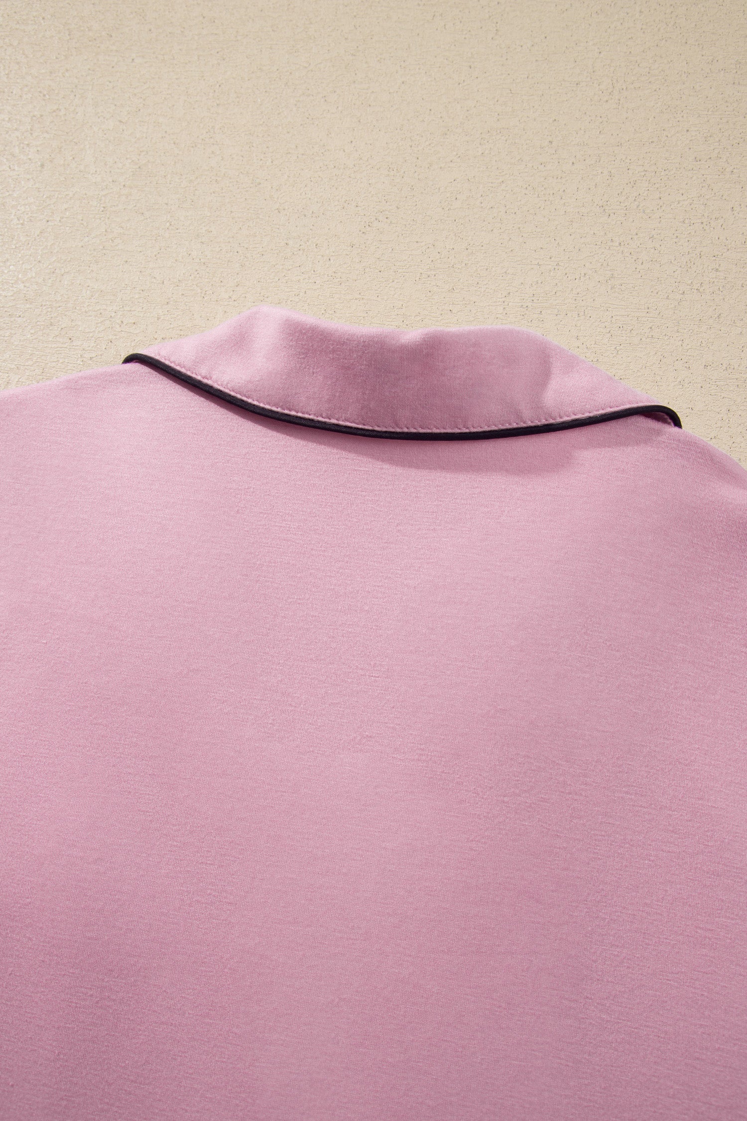 A close-up of a light pink garment, likely a sweatshirt or jacket, showing the back of the neck with a thin black piping trim against a textured beige background.