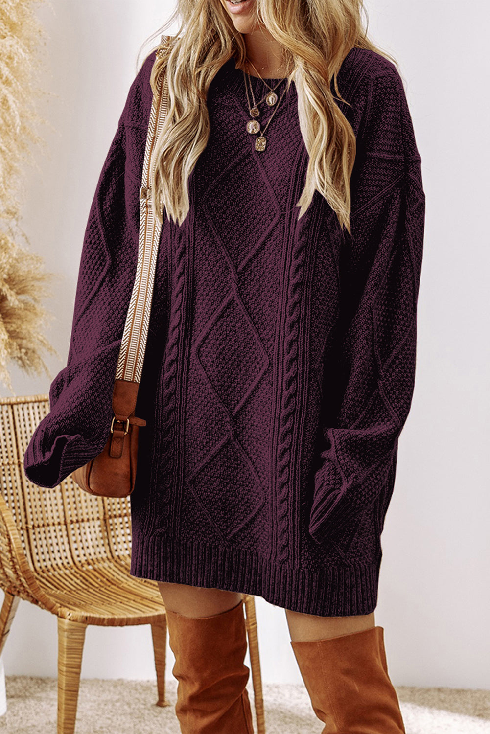 A woman wearing an oversized dark purple cable-knit sweater, styled with a brown crossbody bag and brown over-the-knee boots, standing next to a wicker chair.