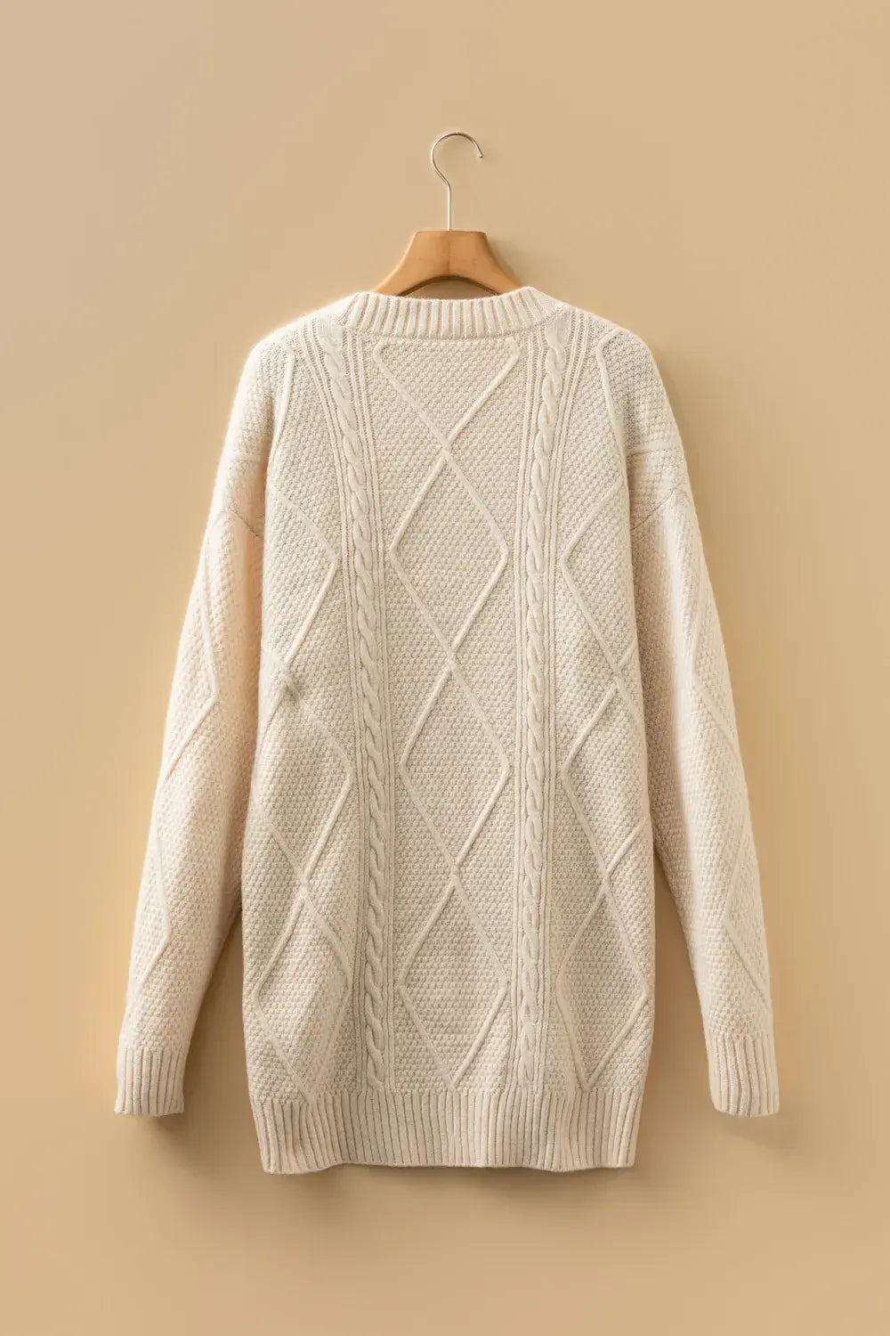 An off-white cable-knit sweater hanging on a wooden hanger against a solid beige background.