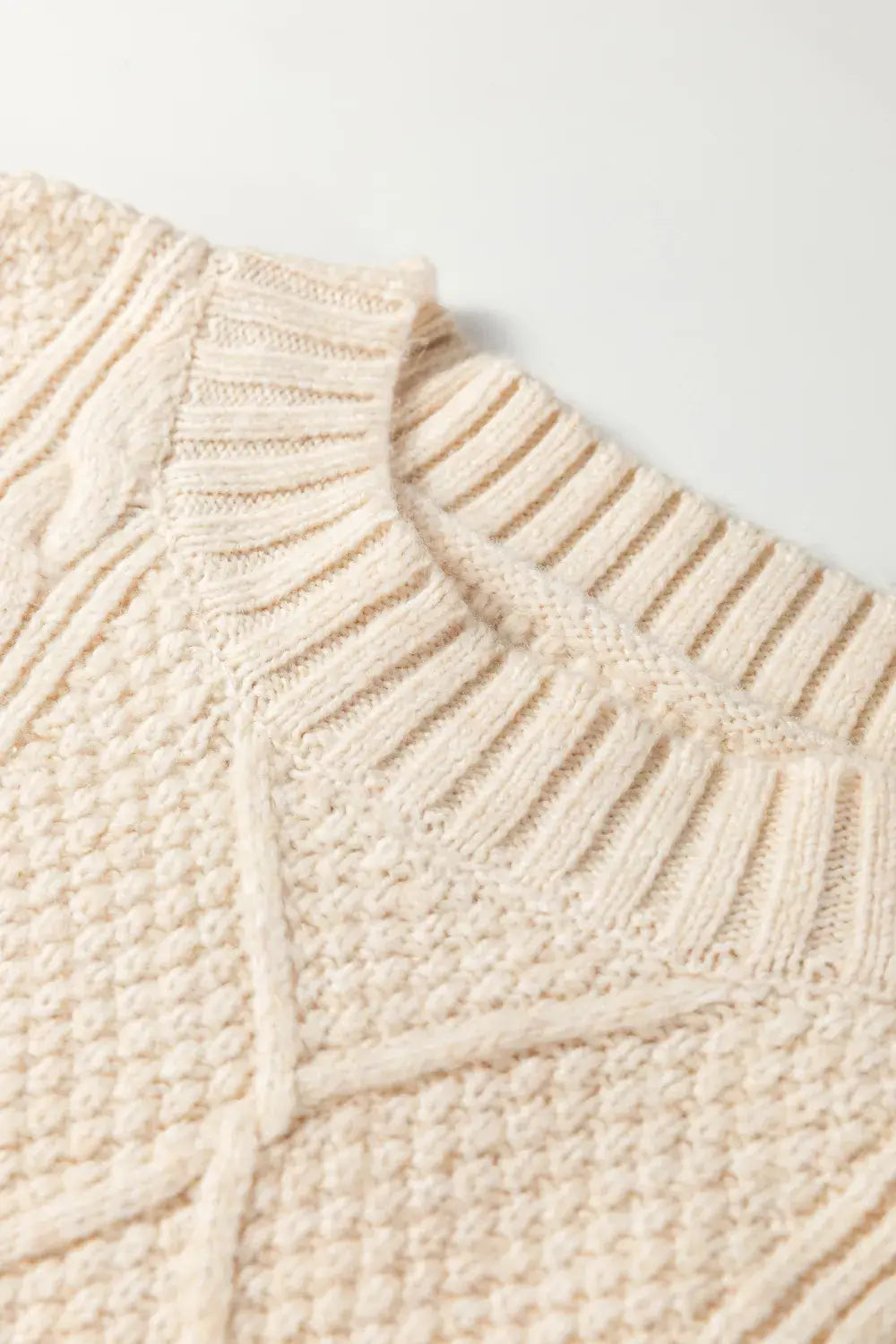 Close-up of a cream-colored knit sweater showing the textured cable and ribbed patterns of the collar and upper chest.