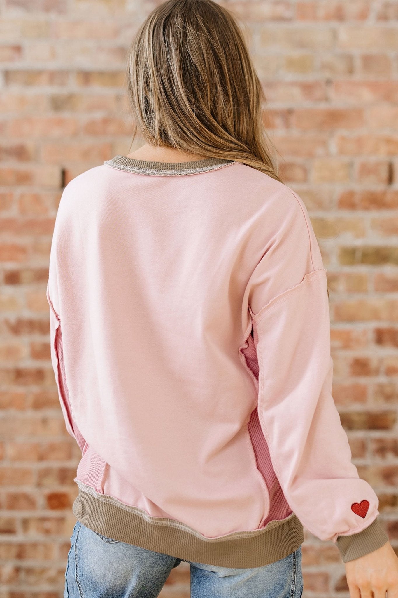Person wearing a pink bomber jacket with a heart on the sleeve against a brick wall.