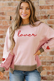 Woman wearing a pink sweatshirt with 'lover' text and heart designs, standing against a brick wall.