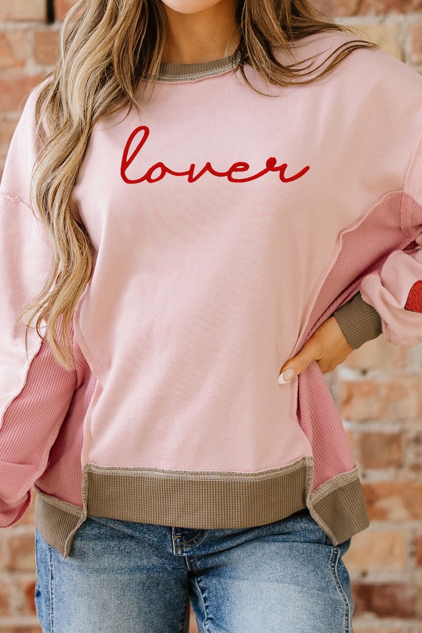 Person wearing a pink sweatshirt with 'lover' printed on it, standing against a brick wall.