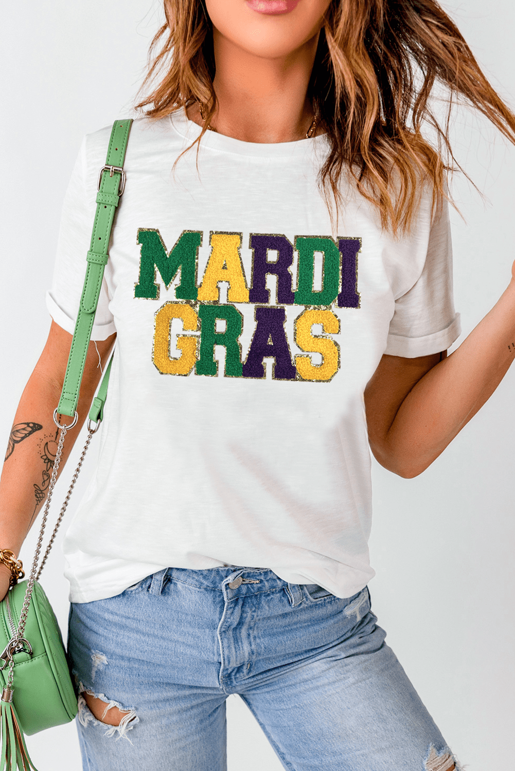 Person wearing a white t-shirt with 'MARDI GRAS' in colorful letters, paired with blue jeans and a green handbag.
