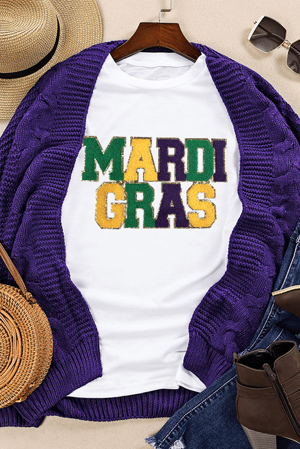 White t-shirt with 'MARDI GRAS' text in colorful letters, purple cardigan, straw bag, and sunglasses on a neutral background.