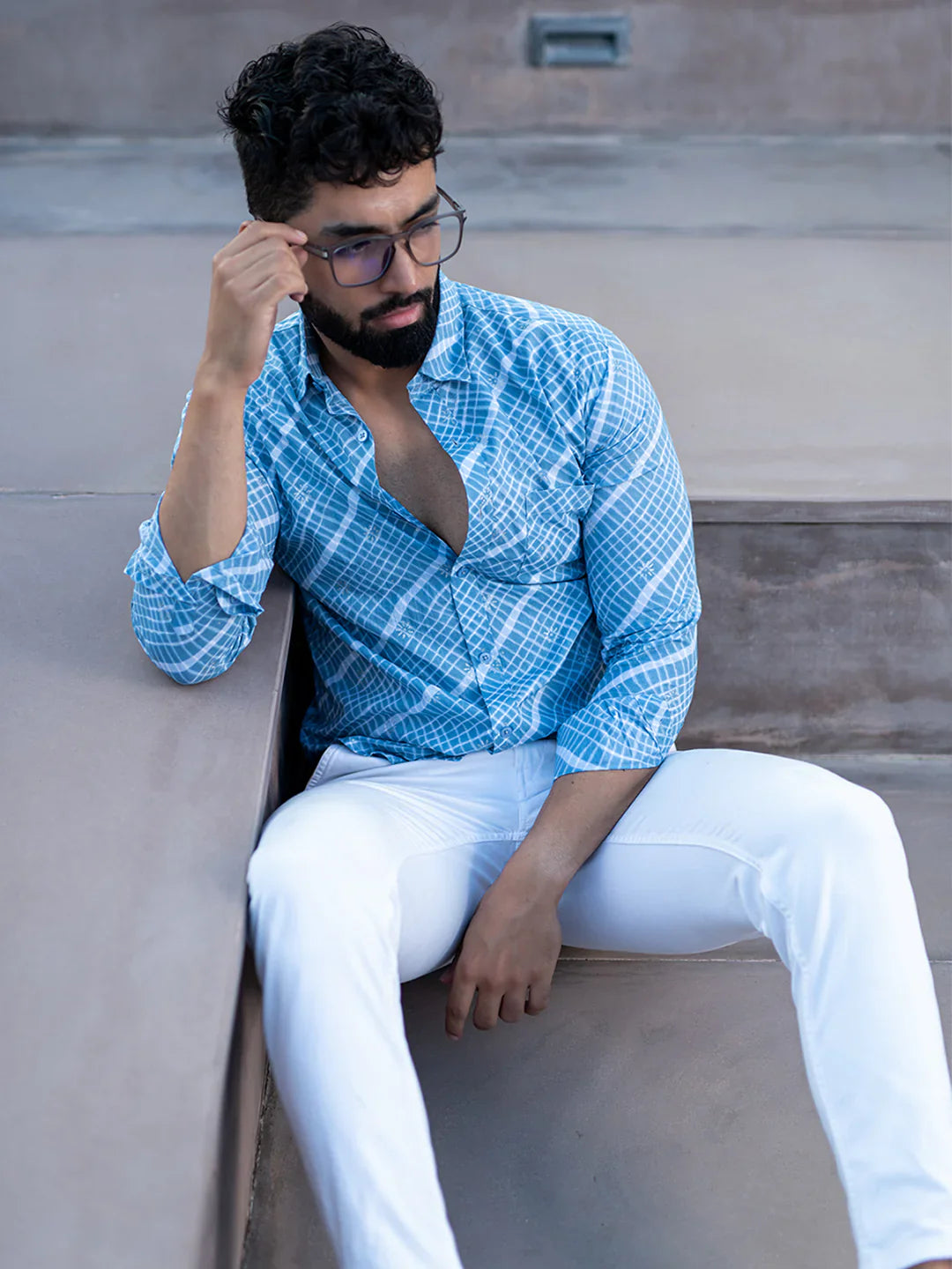 Man wearing a blue patterned shirt and white pants sitting on a bench.