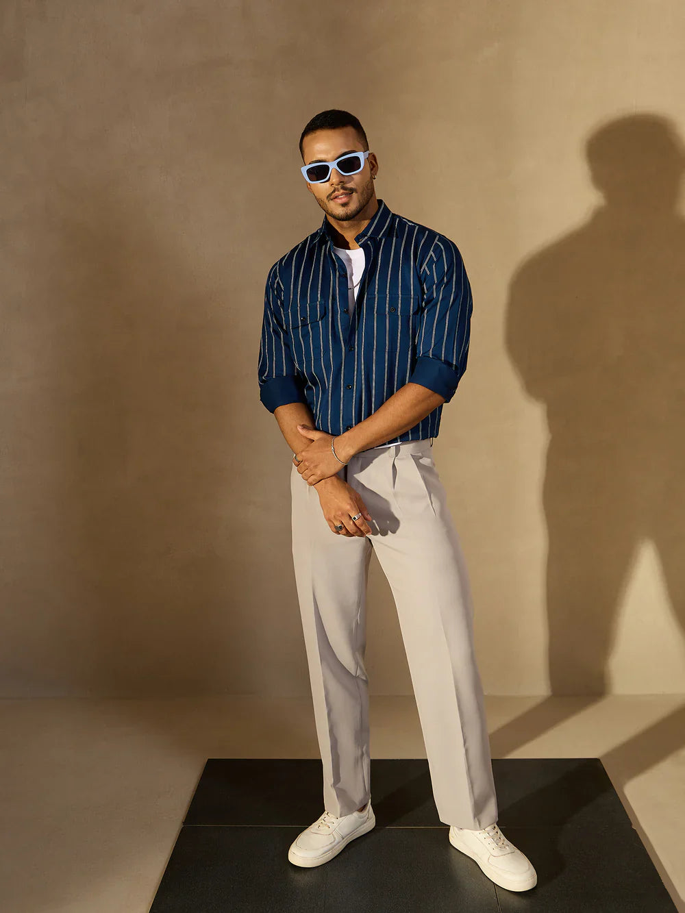 Man wearing a blue striped shirt, white pants, and sunglasses against a beige wall.