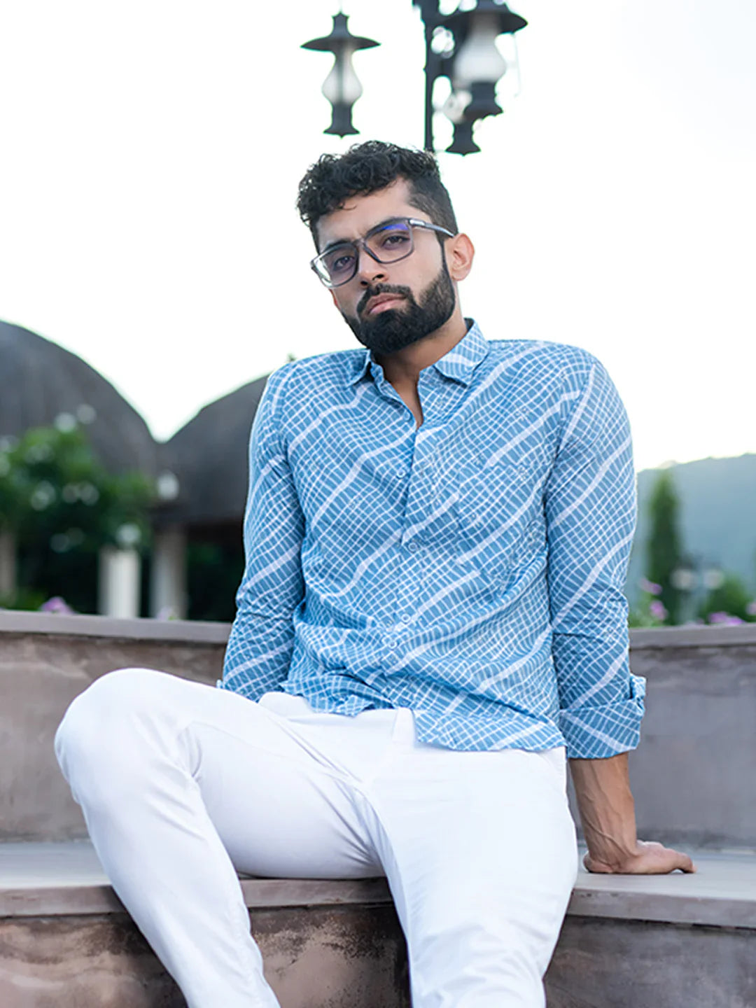 Man wearing a blue patterned shirt and white pants sitting on a ledge with a scenic background.