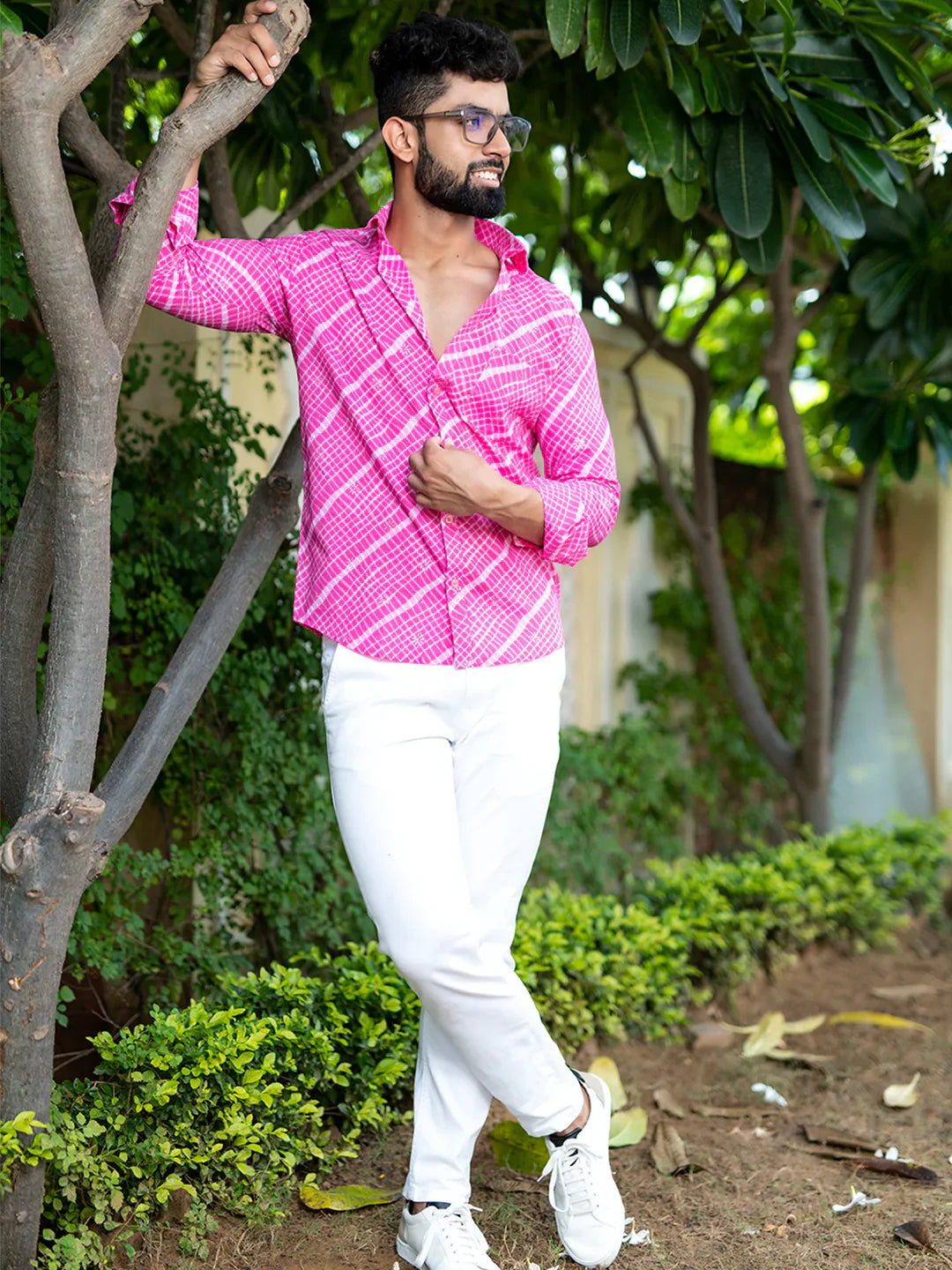 Man wearing a pink patterned shirt and white pants standing outdoors among trees.
