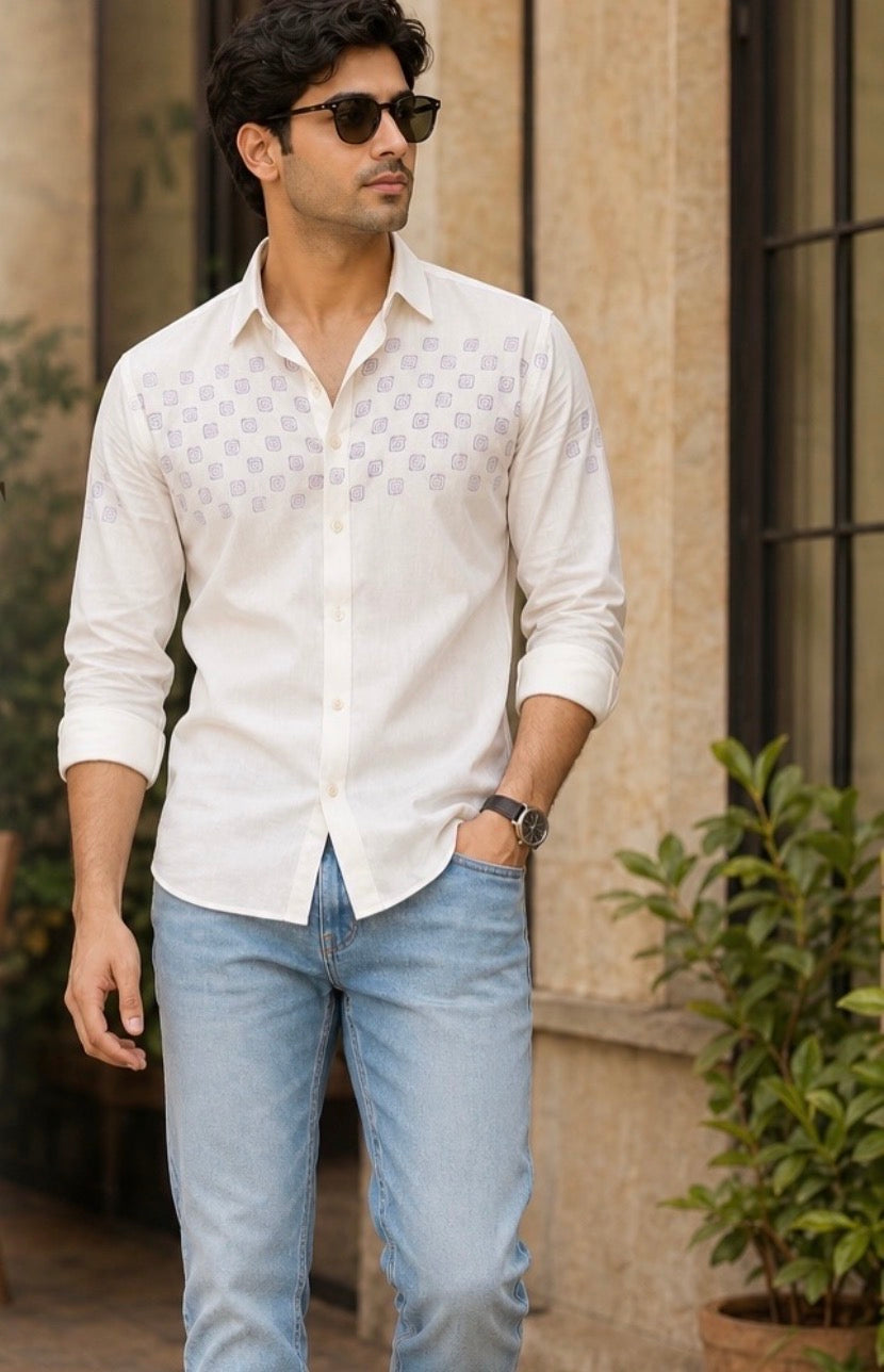 Man wearing a white patterned shirt and light blue jeans standing outdoors.