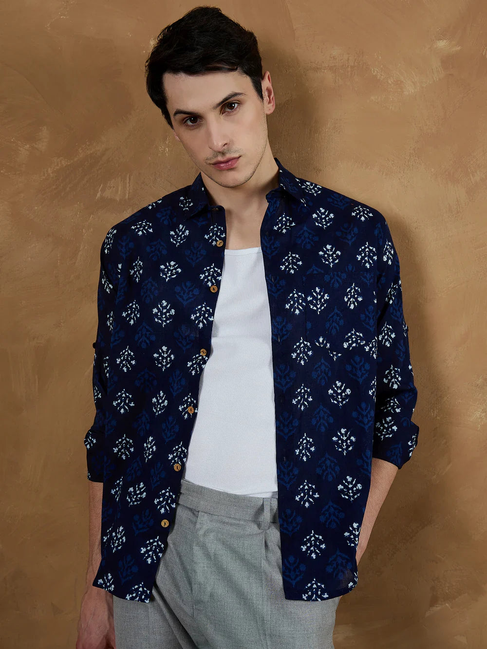 Man wearing a navy patterned shirt over a white tank top with gray pants against a brown background
