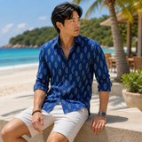 Man wearing a blue patterned shirt and white shorts sitting on a beach.