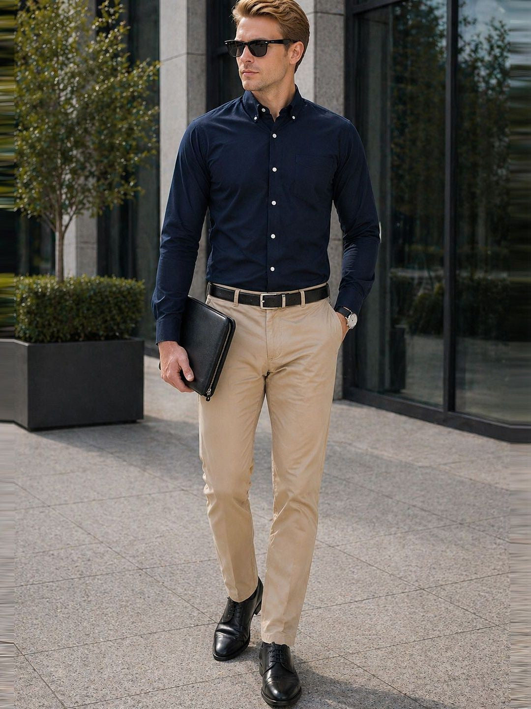 Man in navy shirt and beige pants holding a black folder outdoors.