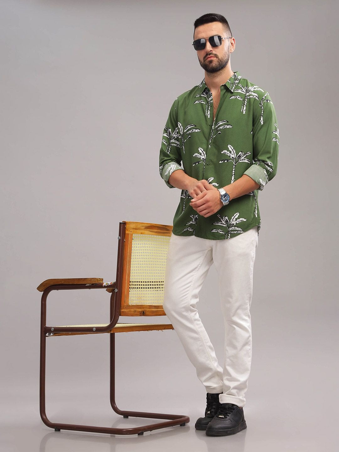 Man wearing a green patterned shirt and white pants standing next to a wooden chair on a gray background