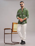 Man wearing a green patterned shirt and white pants standing next to a wooden chair on a gray background