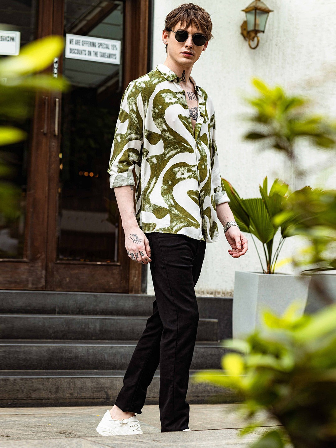 Person wearing a patterned shirt and sunglasses outdoors with plants and a building in the background