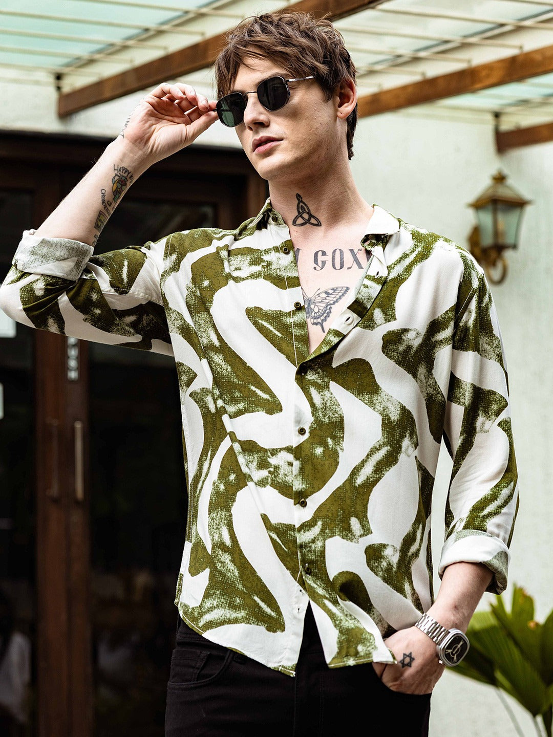 Person wearing a patterned shirt with tattoos and sunglasses outdoors.