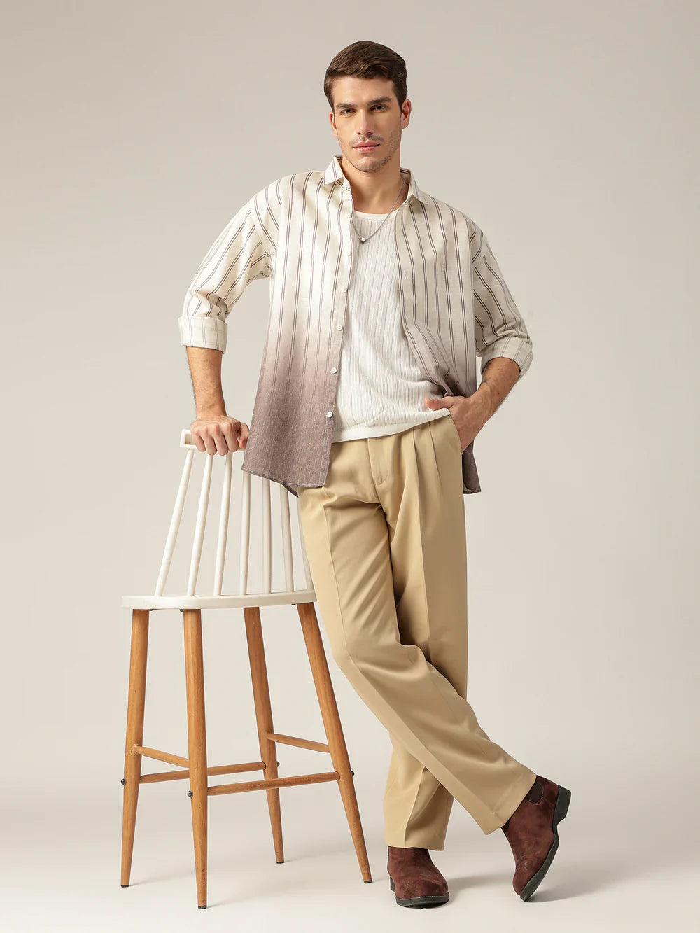 Man wearing a striped shirt and beige pants standing next to a wooden chair on a plain background