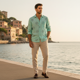 Man in a light green shirt and beige pants standing by a waterfront with buildings in the background.
