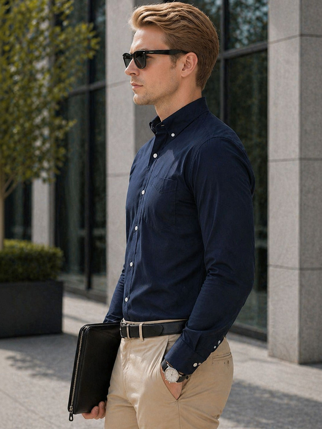 Man in navy shirt and beige pants holding a black folder, standing outdoors.