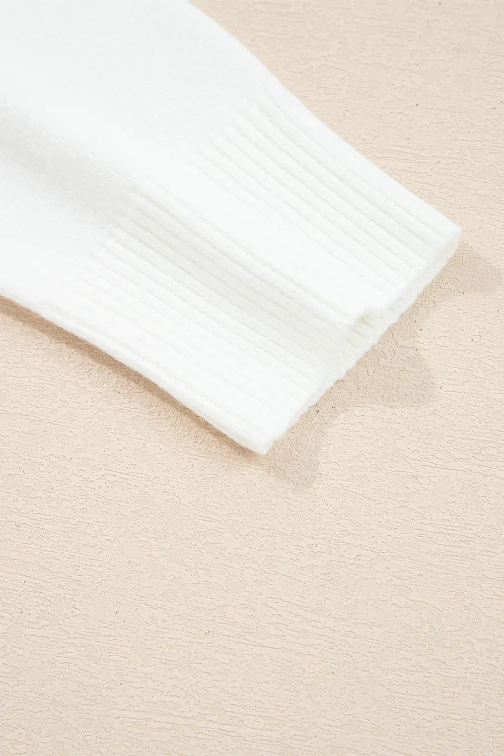 Close-up of a white ribbed knit sleeve or cuff laid diagonally on a light beige textured surface.
