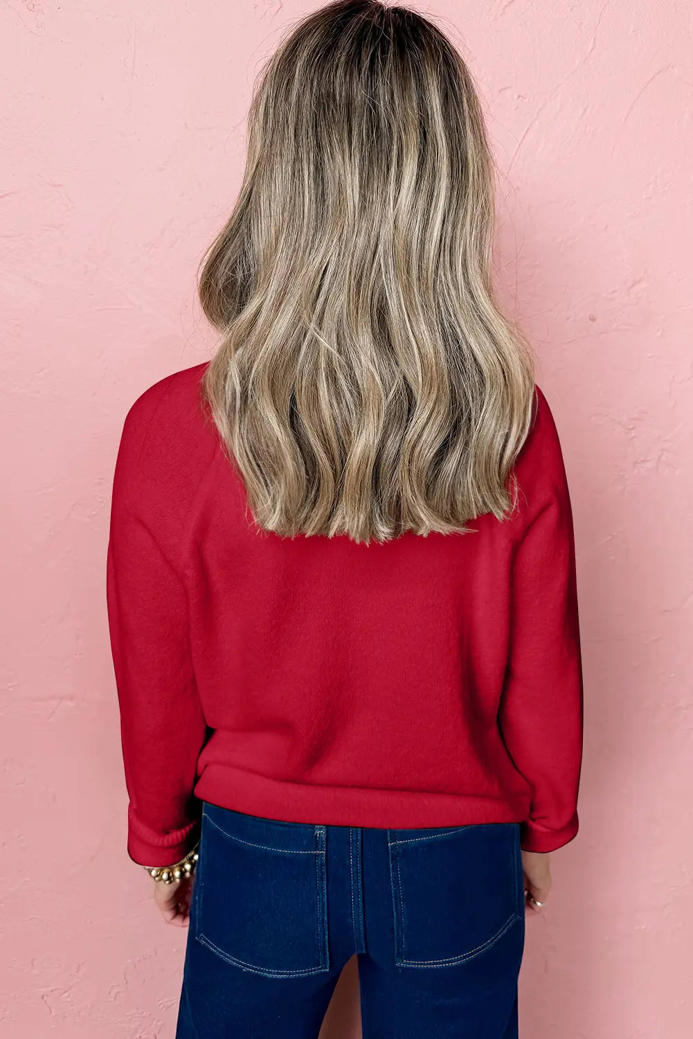 A woman viewed from behind wearing a bright red long-sleeve sweatshirt and dark blue jeans, standing against a textured light pink wall.