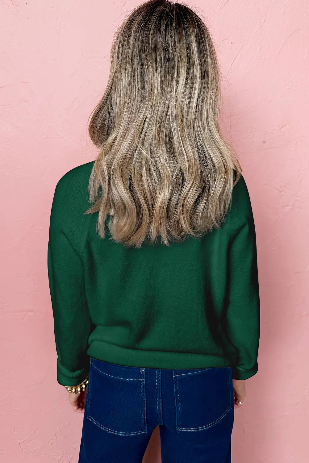 Person wearing a green sweater and blue jeans against a pink wall