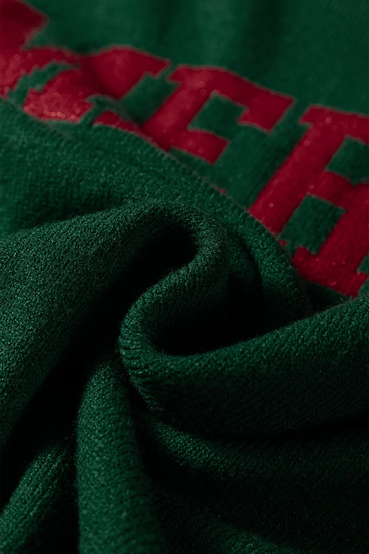 Close-up of dark green fabric with a red embroidered letter detail, showing the texture of the material.