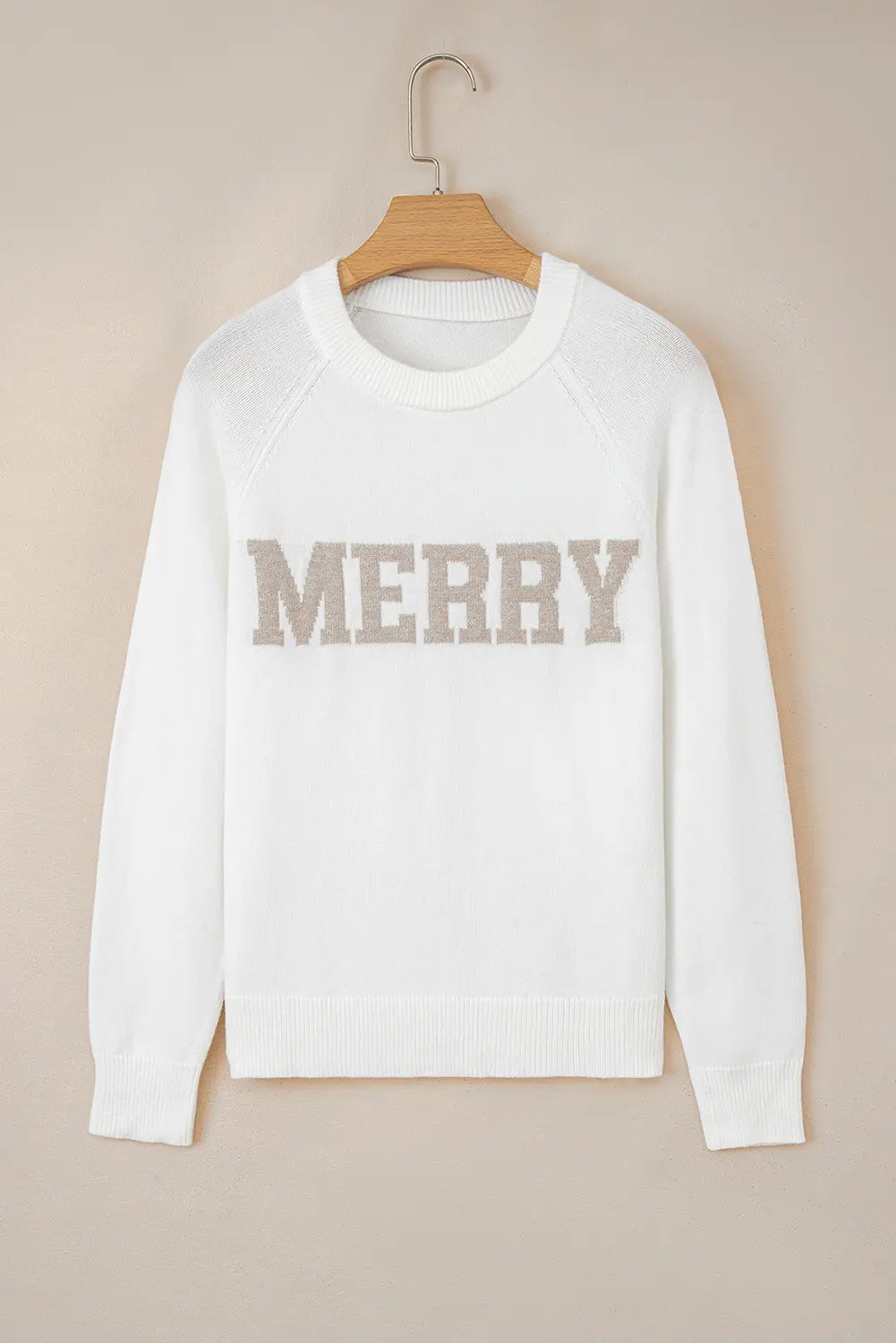 A white crewneck knit sweater hanging on a wooden hanger against a warm beige background. The word 'MERRY' is knitted across the chest in a textured taupe-brown lettering.