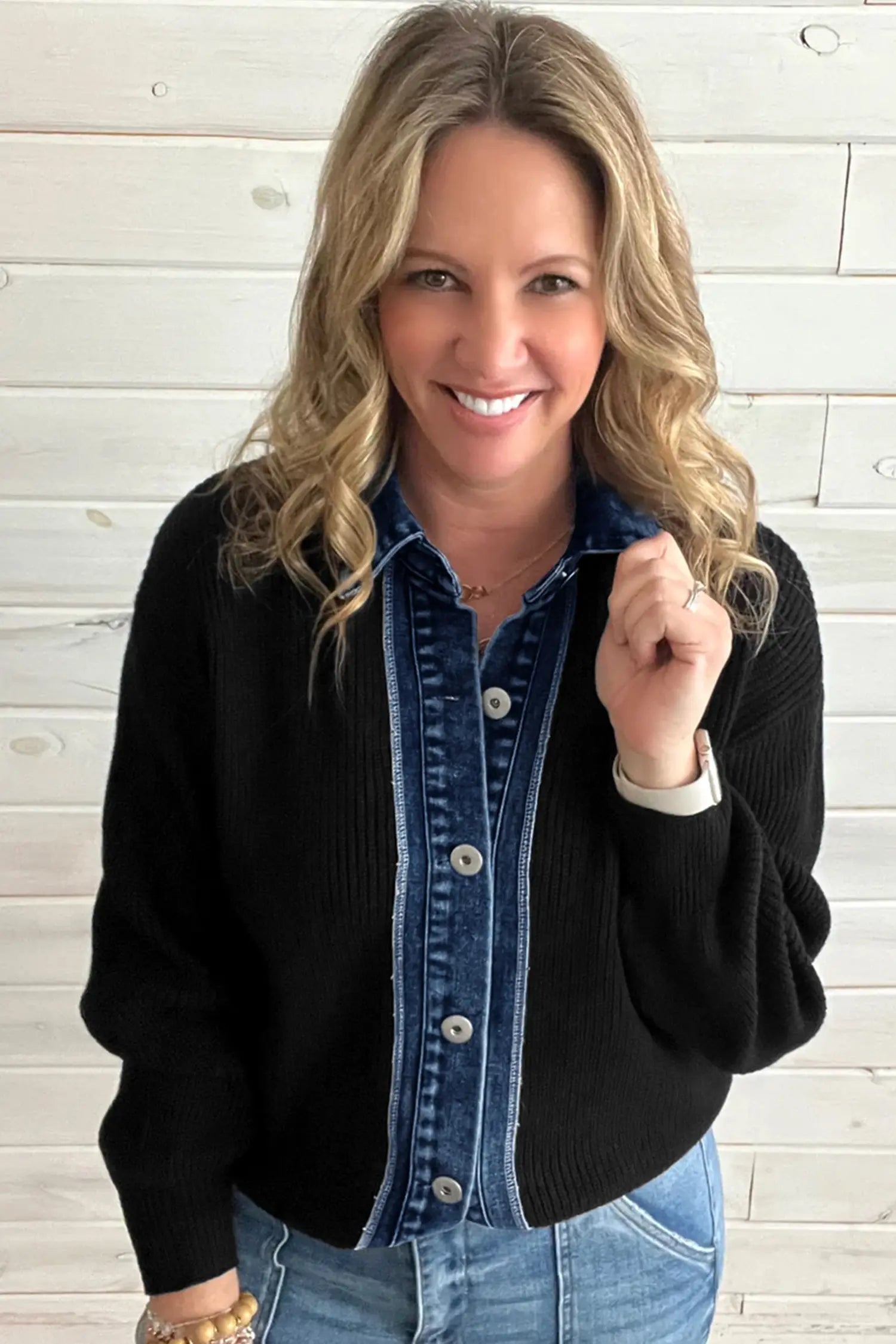A smiling blonde woman wearing a black ribbed knit cardigan over a blue denim button-up shirt, paired with light blue jeans, standing against a white wood plank wall.