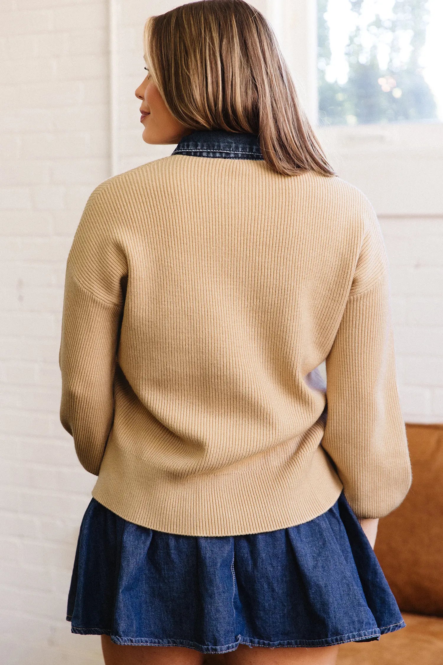 A woman shown from the back wearing a beige ribbed knit sweater over a dark blue denim shirt and a matching denim pleated skirt, standing in front of a white brick wall.