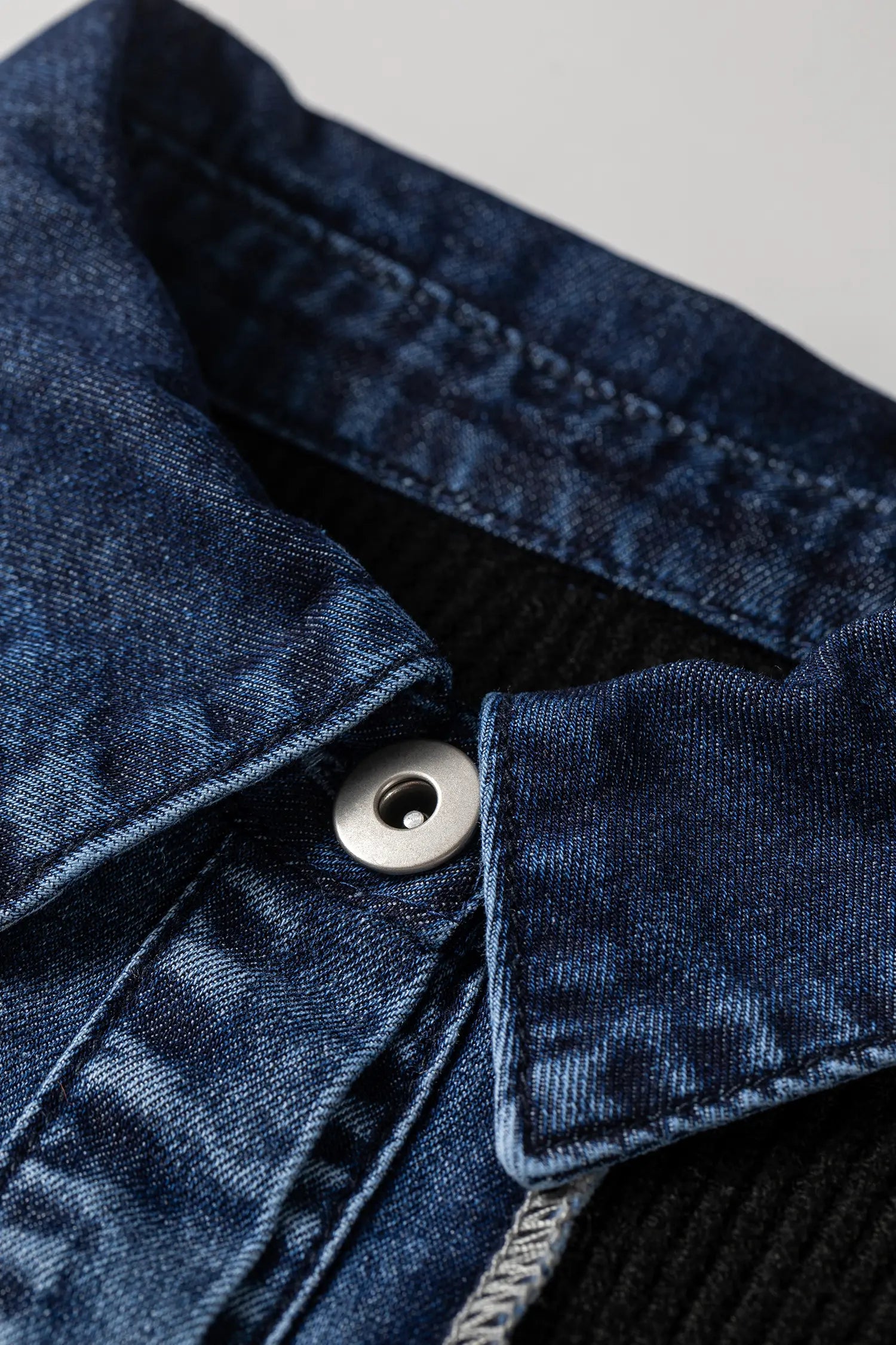 Close-up detail of a dark blue denim jacket collar with a silver snap button, showing the textured denim weave and a black knit layer underneath.