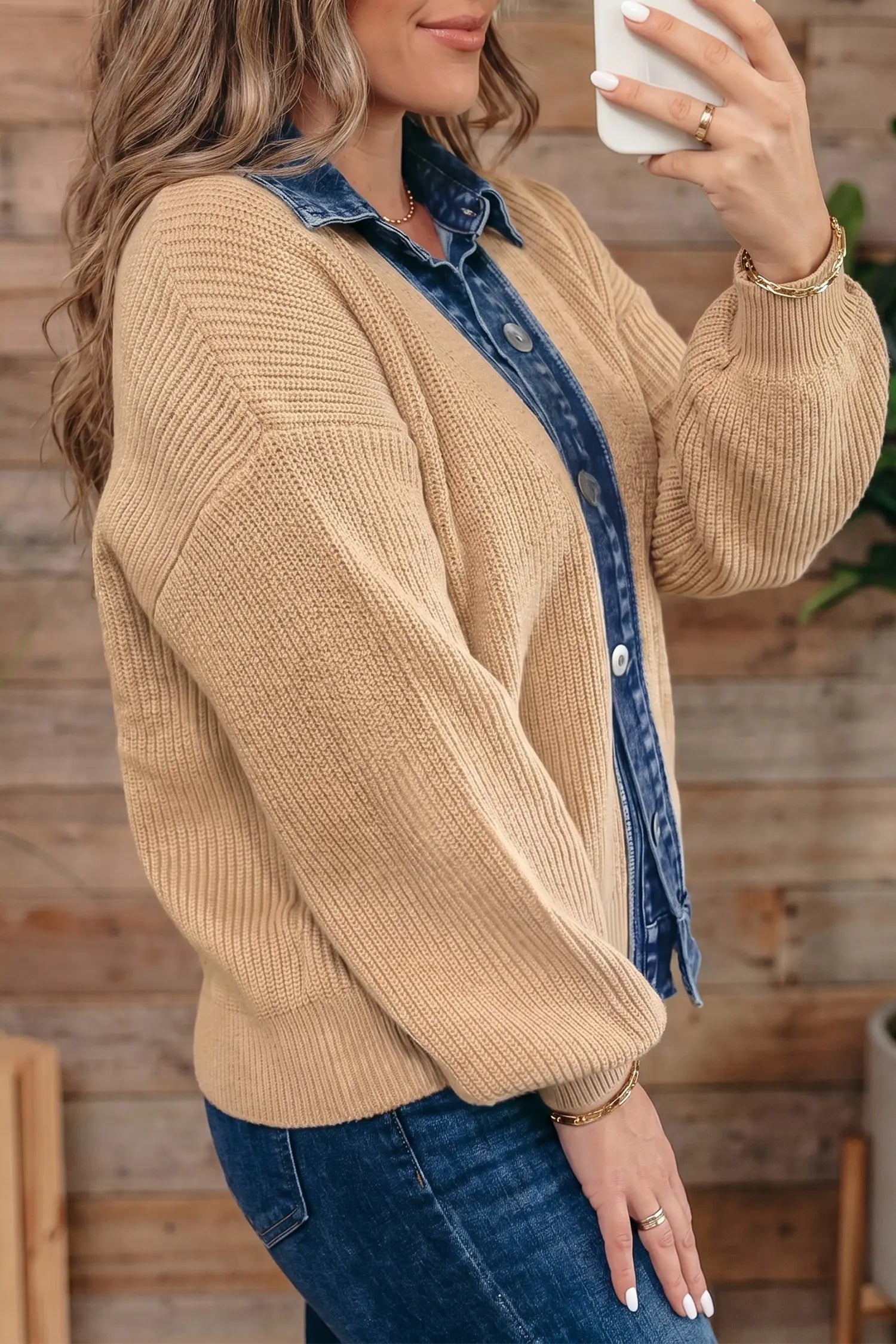 A woman wearing a beige ribbed knit cardigan over a blue denim button-up shirt and blue jeans, holding a white phone in front of her face. The background is a rustic wooden wall.