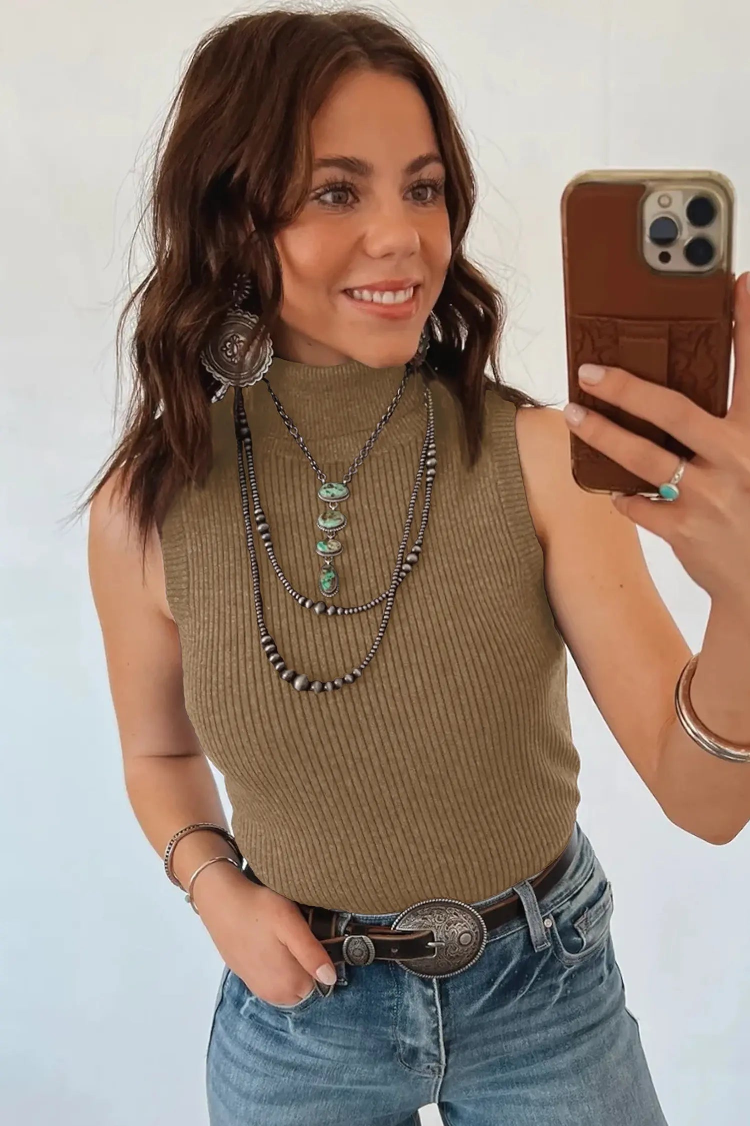 Woman taking a mirror selfie wearing a brown sleeveless top and blue jeans, with layered necklaces and bracelets.