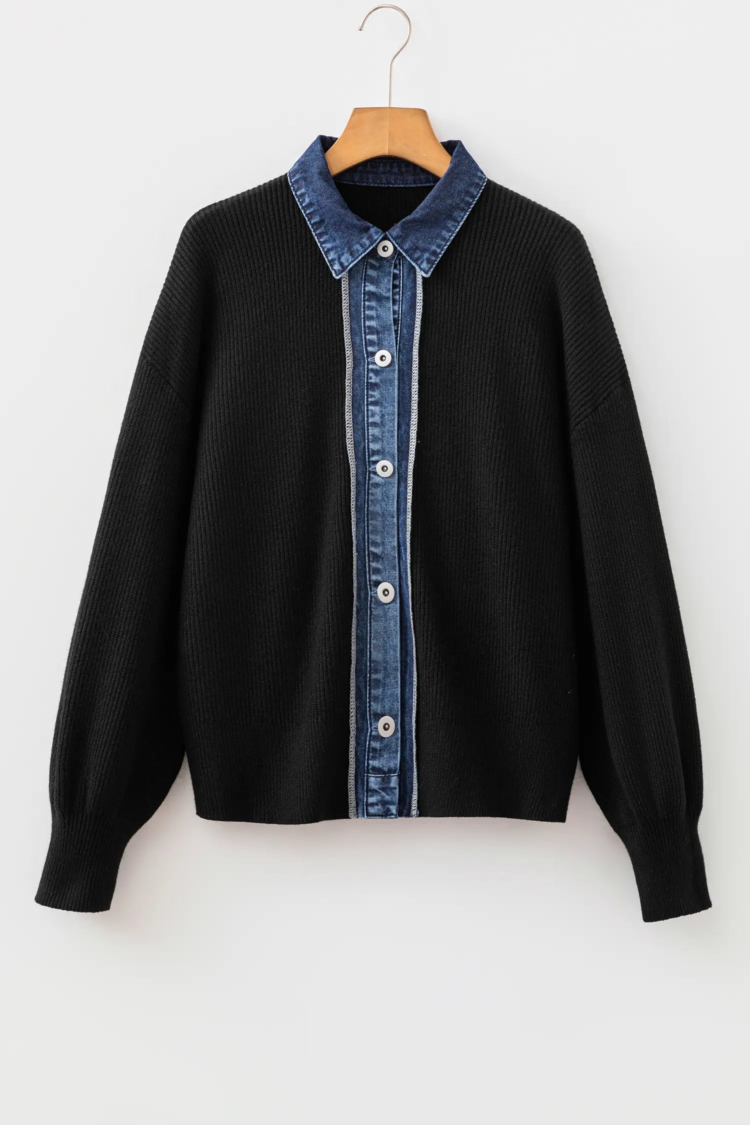 A black ribbed knit cardigan sweater with a blue denim button-up shirt collar and front placket, hanging on a wooden hanger against a plain white wall.