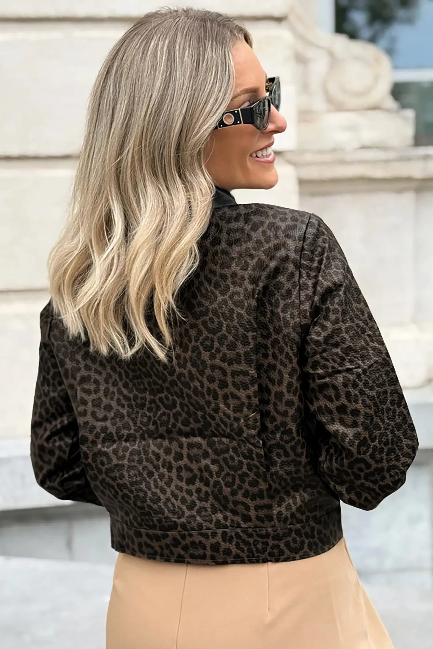 A woman with blonde hair and sunglasses, viewed from the back, wearing a dark brown and black leopard print bomber jacket and beige pants.