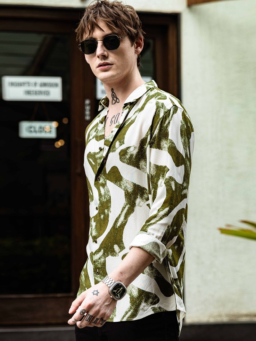 Person wearing a green and white patterned shirt with sunglasses and a tattoo.